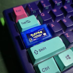 May include: A custom artisan keycap shaped like a blue translucent Game Boy Advance Pok&eacute;mon Sapphire Version game cartridge, installed on a mechanical keyboard with purple and teal keys, including a pink Esc key and a teal Tab key.