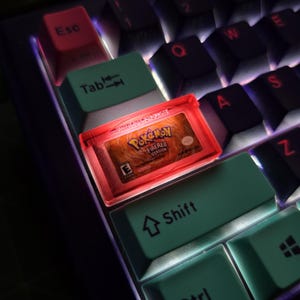 May include: A custom mechanical keyboard keycap featuring a miniature replica of a Pokemon FireRed Version game cartridge. The keycap is translucent red and illuminated from within, displaying the game logo and E rating clearly on the surface.