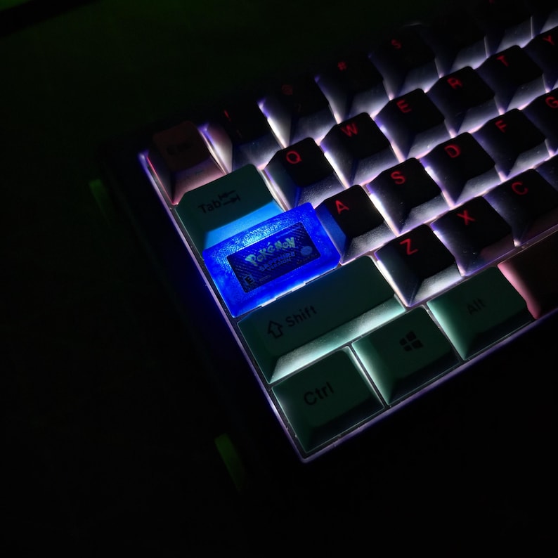 May include: A mechanical keyboard featuring a custom translucent blue keycap shaped like a Game Boy Advance game cartridge, specifically labeled Pok&eacute;mon Sapphire Version, illuminated by internal backlighting among other light-colored keys.