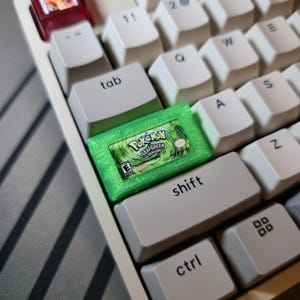 May include: A custom mechanical keyboard keycap shaped like a translucent green Game Boy Advance game cartridge. The keycap features a detailed label for Pok&eacute;mon LeafGreen Version, including the game title, the E rating symbol, and Nintendo logo.