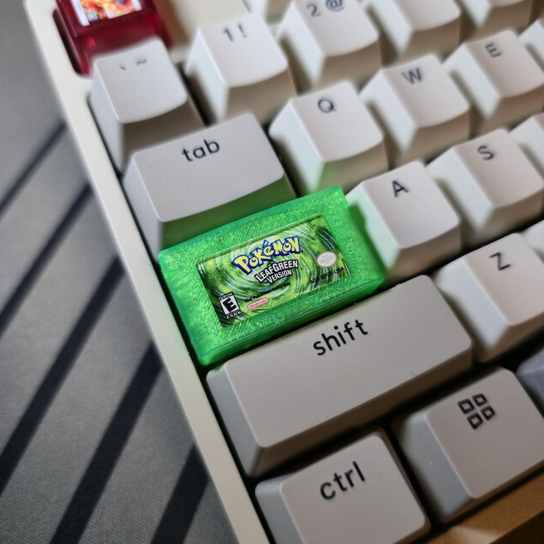 May include: A custom mechanical keyboard keycap shaped like a translucent green Game Boy Advance game cartridge. The keycap features a detailed label for Pok&eacute;mon LeafGreen Version, including the game title, the E rating symbol, and Nintendo logo.