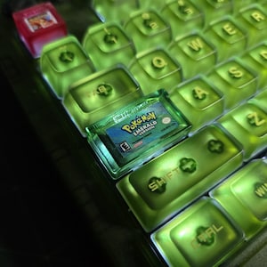 May include: A mechanical keyboard with translucent lime green keycaps and gold lettering. A Game Boy Advance Pok&eacute;mon Emerald Version game cartridge is placed on the keyboard, and a small red translucent keycap is visible in the top left corner.