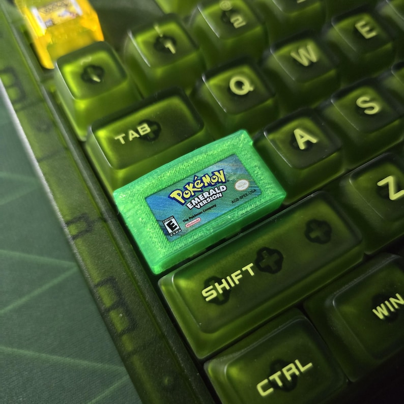 May include: A custom mechanical keyboard keycap shaped like a miniature Pokemon Emerald Version game cartridge, featuring a green translucent housing and a detailed label, installed on a dark green translucent mechanical keyboard.