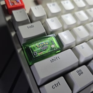 May include: A custom artisan keycap shaped like a translucent green Game Boy Advance Pok&eacute;mon LeafGreen Version game cartridge, positioned on a white mechanical keyboard next to the tab and shift keys, with a small red cartridge visible above.