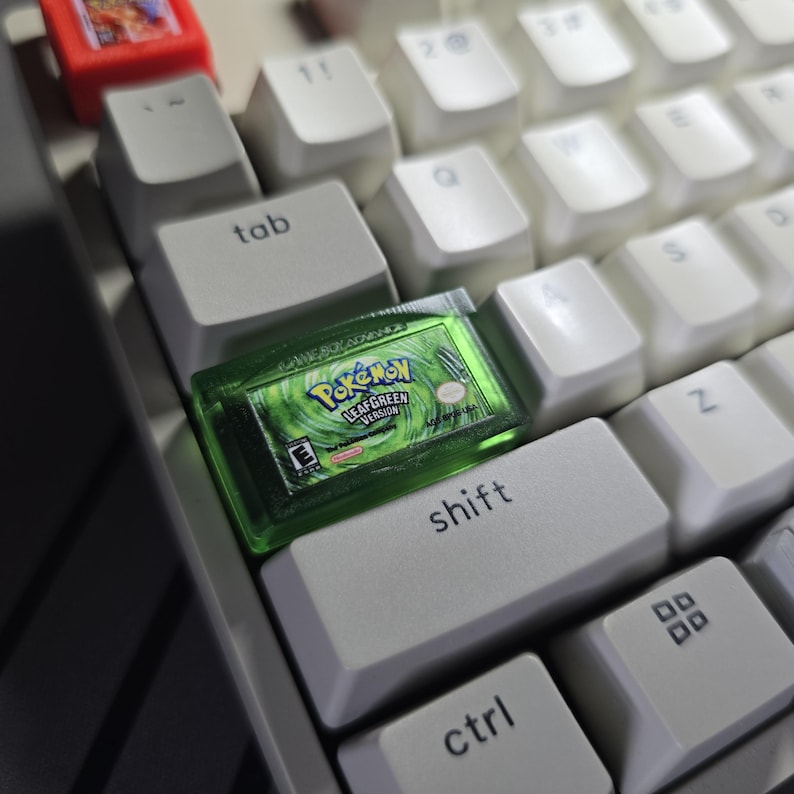 May include: A custom artisan keycap shaped like a translucent green Game Boy Advance Pok&eacute;mon LeafGreen Version game cartridge, positioned on a white mechanical keyboard next to the tab and shift keys, with a small red cartridge visible above.