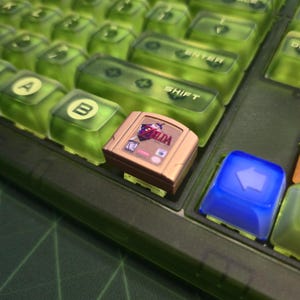 May include: A close-up of a mechanical keyboard with translucent green keycaps. A gold-colored Nintendo 64 game cartridge is placed on the keyboard. A blue keycap with a white arrow is also visible.
