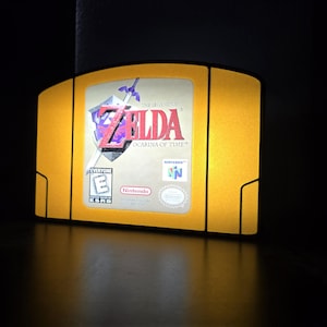 May include: A yellow-colored lamp shaped like a Nintendo 64 game cartridge. The label reads "The Legend of Zelda: Ocarina of Time" with the Nintendo logo and an ESRB rating. The lamp is illuminated, casting a warm glow.