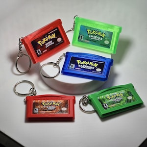 May include: A collection of five miniature game cartridge keychains featuring Pokémon Ruby, Emerald, Sapphire, FireRed, and LeafGreen versions. Each plastic cartridge includes a metal keyring and displays the official game title, logo, and ESRB rating.