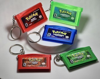 May include: A collection of five miniature game cartridge keychains featuring Pokémon Ruby, Emerald, Sapphire, FireRed, and LeafGreen versions. Each plastic cartridge includes a metal keyring and displays the official game title, logo, and ESRB rating.