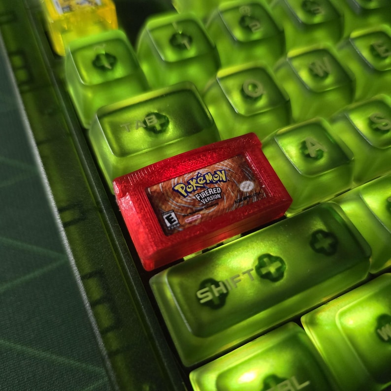 May include: A translucent red custom keycap shaped like a Pokemon FireRed game cartridge, featuring the game title and logo, positioned on a mechanical keyboard with translucent lime green keycaps that have black lettering on the surface.