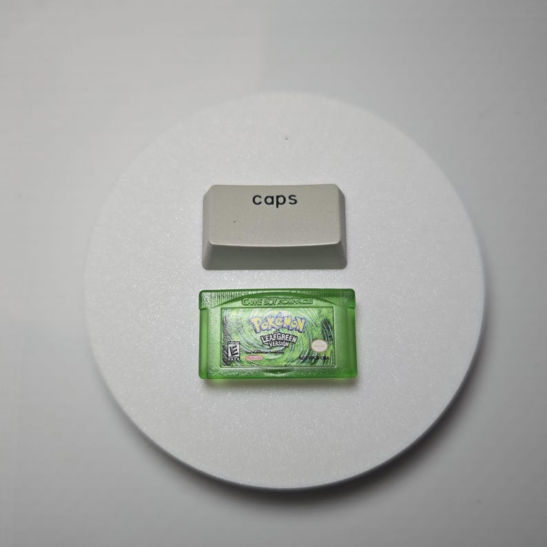 May include: A light gray keyboard keycap with the word caps printed in black, positioned above a translucent green Game Boy Advance video game cartridge labeled Pokemon LeafGreen Version with an E rating symbol and Nintendo logo.