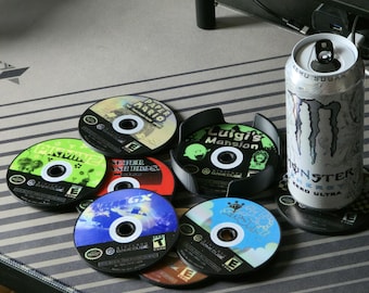 May include: A collection of Nintendo GameCube game discs including Paper Mario, Pikmin 2, Super Smash Bros. Melee, F-Zero GX, Luigi's Mansion, and Super Mario Sunshine, alongside a silver Monster Energy Zero Ultra can on a gray striped mat.