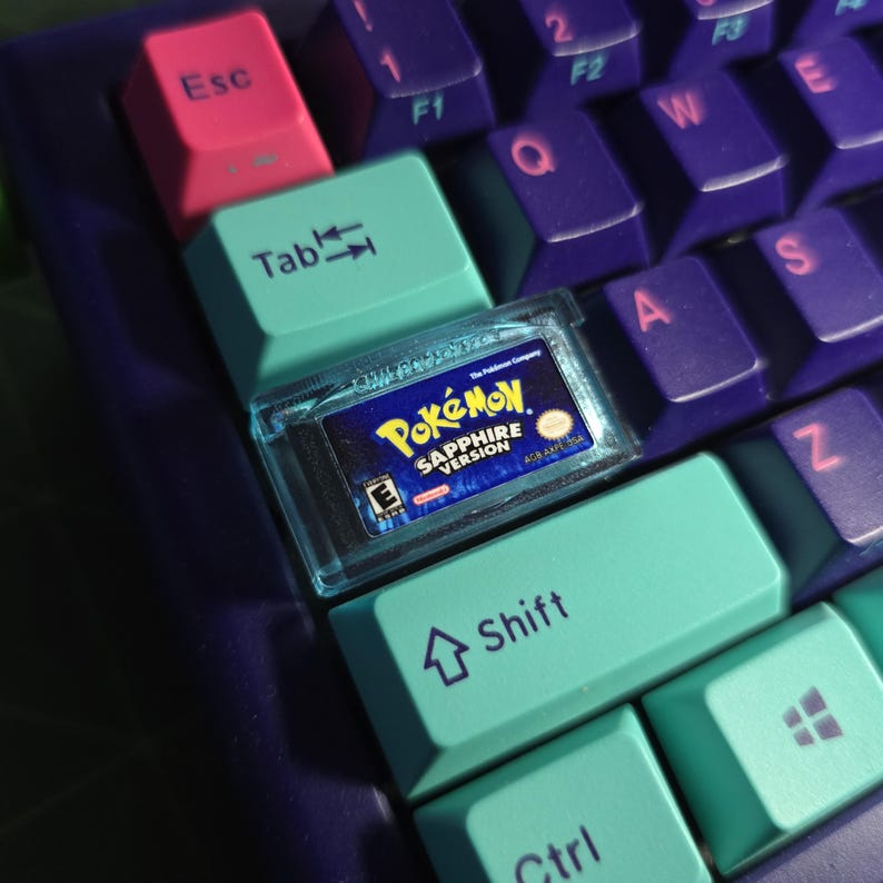 May include: A custom mechanical keyboard keycap shaped like a miniature Pokemon Sapphire Version game cartridge. The keycap is translucent blue and sits on a keyboard with purple, pink, and teal keys featuring printed text and symbols.