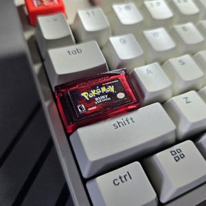 May include: A custom mechanical keyboard keycap shaped like a translucent red Pokemon Ruby Version game cartridge. The keycap features the official game logo, the text Pokemon Ruby Version, and an E rating symbol printed on the front surface.
