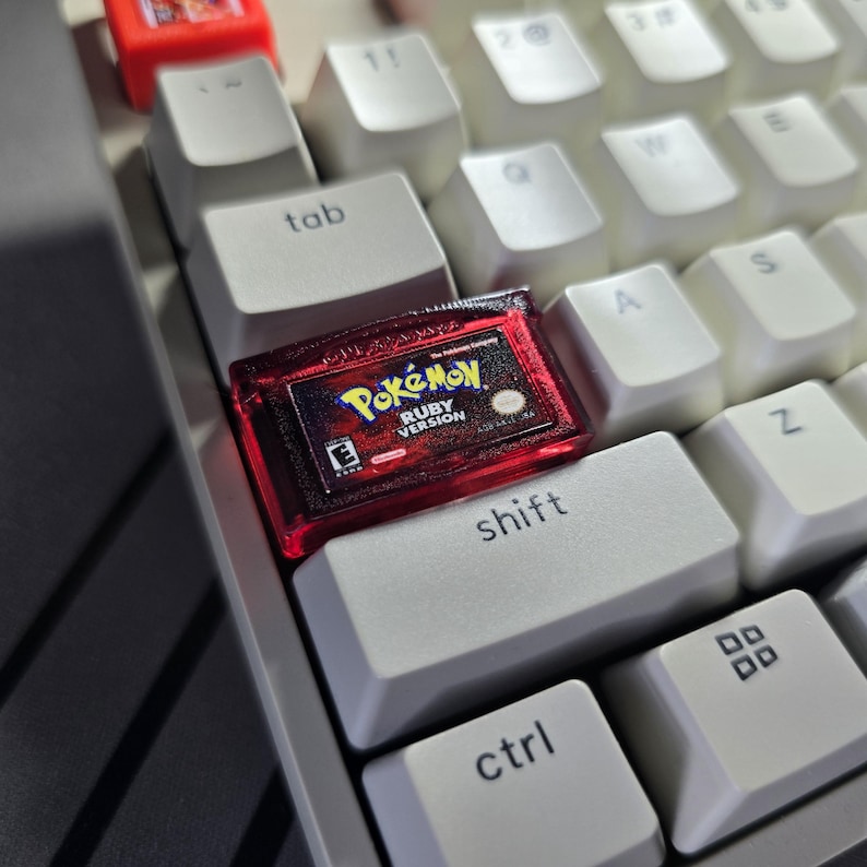 May include: A custom mechanical keyboard keycap shaped like a translucent red Pokemon Ruby Version game cartridge. The keycap features the official game logo, the text Pokemon Ruby Version, and an E rating symbol printed on the front surface.