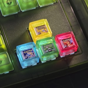May include: Four translucent, square mechanical keyboard keycaps shaped like Game Boy cartridges. Each keycap features a miniature Pokémon game label, including Pikachu, Venusaur, Blastoise, and Charizard, with the Game Boy logo embossed on the top.
