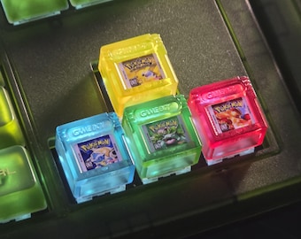 May include: Four translucent, square mechanical keyboard keycaps shaped like Game Boy cartridges. Each keycap features a miniature Pokémon game label, including Pikachu, Venusaur, Blastoise, and Charizard, with the Game Boy logo embossed on the top.