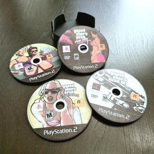 May include: Four circular PlayStation 2 game discs featuring Grand Theft Auto titles including Vice City, San Andreas, and Grand Theft Auto III. Each disc displays the PlayStation 2 logo, the ESRB M rating, and specific character or weapon artwork.
