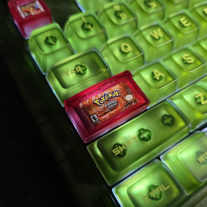 May include: A mechanical keyboard featuring translucent lime green keycaps with gold lettering. A custom red keycap shaped like a Nintendo Game Boy Advance cartridge is installed, featuring a label that reads Pok&eacute;mon FireRed Version.
