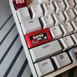 May include: A custom mechanical keyboard keycap shaped like a miniature red translucent Pokemon Ruby Version game cartridge, featuring the game logo and E rating, installed on a white keyboard among standard white keys with black lettering.
