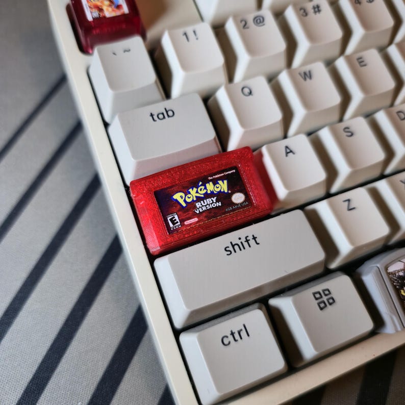 May include: A custom mechanical keyboard keycap shaped like a miniature red translucent Pokemon Ruby Version game cartridge, featuring the game logo and E rating, installed on a white keyboard among standard white keys with black lettering.