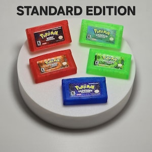 May include: Five rectangular video game cartridges labeled STANDARD EDITION, including Pokemon Ruby, Emerald, FireRed, LeafGreen, and Sapphire versions. The cartridges are translucent red, green, and blue plastic with detailed game title labels.