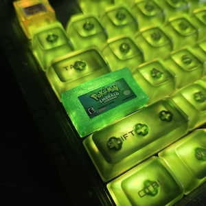 May include: A custom mechanical keyboard keycap shaped like a Nintendo Game Boy Advance game cartridge, specifically Pok&eacute;mon Emerald Version, integrated into a set of translucent neon green backlit keycaps with visible labels like TAB and SHIFT.