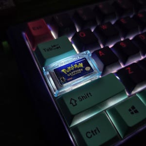 May include: A custom mechanical keyboard keycap featuring a miniature, translucent blue replica of a Pokemon Sapphire Version game cartridge. The keycap is positioned between light blue Tab and Shift keys on a backlit keyboard with dark keys.