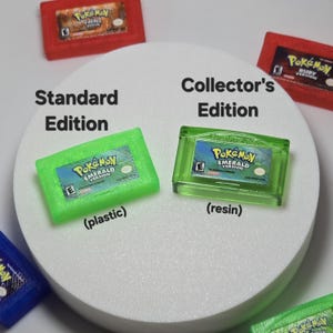 May include: Two green Pokemon Emerald Version game cartridges labeled Standard Edition in plastic and Collector's Edition in resin, surrounded by other red and blue game cartridges on a white circular surface with text labels.