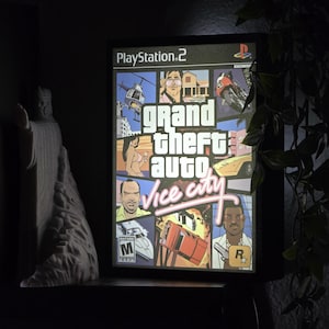 May include: A rectangular, illuminated display featuring the Grand Theft Auto Vice City video game cover art for PlayStation 2. The graphic includes a collage of characters, a helicopter, a sports car, and a boat, with the game title in pink text.