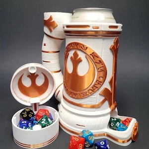Puede incluir: Una torre de dados y un contenedor de almacenamiento blancos y bronce con temática de Star Wars. La torre tiene un diseño cilíndrico con el símbolo y el texto de la Alianza Rebelde. Un contenedor separado contiene dados de varios colores.