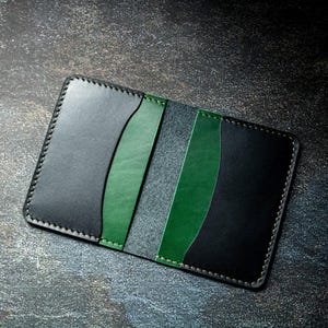 May include: A black leather cardholder with green and grey panels. The wallet is hand-stitched with white thread and has a bi-fold design. The cardholder is a stylish accessory, with a modern and minimalist aesthetic.
