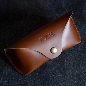 Könnte beinhalten: Ein braunes Lederetui für Brillen mit einem goldenen Knopfverschluss. Das Etui ist mit den Initialen "K.X.H." personalisiert.