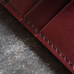May include: Close-up of a burgundy leather wallet with black stitching. The stitching is visible along the edge of the wallet.