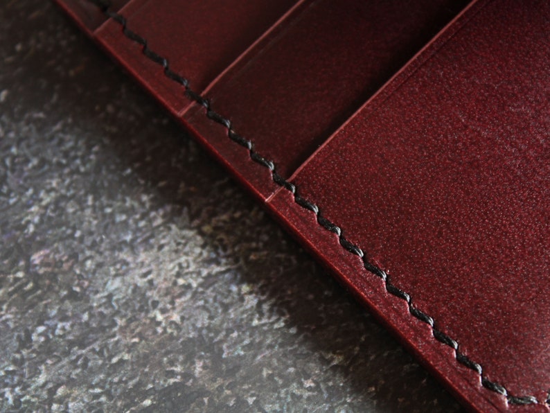 May include: Close-up of a burgundy leather wallet with black stitching. The stitching is visible along the edge of the wallet.
