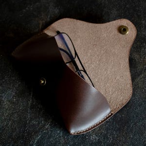 May include: A brown leather glasses case with a flap closure and a brass button. The case is open and a pair of glasses is inside.