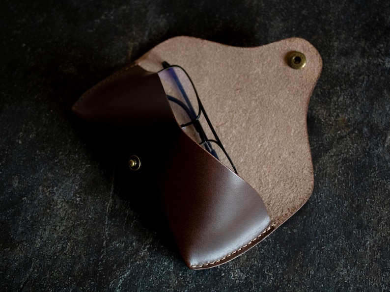 May include: A brown leather glasses case with a flap closure and a brass button. The case is open and a pair of glasses is inside.