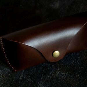 May include: A brown leather glasses case with a gold button closure.