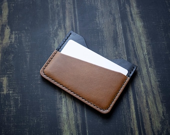 May include: A brown and black leather card holder with a stitched border. The card holder contains white cards. The leather is a rich brown colour, and the edges are black.