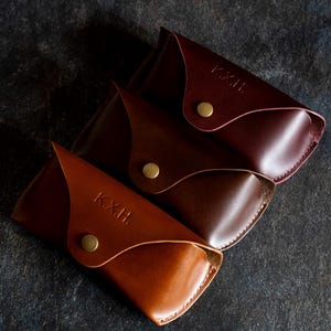 Könnte beinhalten: Drei Lederbrillenetuis, die übereinander gestapelt sind. Das oberste Etui ist burgunderrot, das mittlere Etui ist braun und das unterste Etui ist hellbraun. Jedes Etui hat die Buchstaben "K.X.H." auf der Vorderseite geprägt.