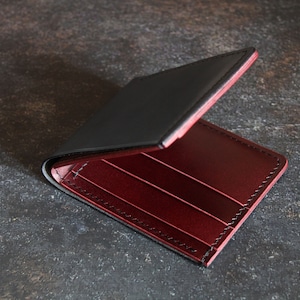 May include: A black leather wallet with a red leather interior. The wallet is open and the interior is visible. The wallet has multiple card slots and a billfold.