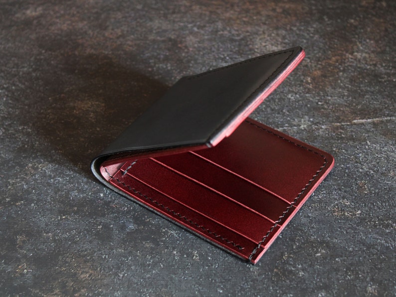 May include: A black leather wallet with a red leather interior. The wallet is open and the interior is visible. The wallet has multiple card slots and a billfold.