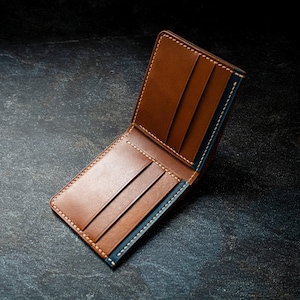 Op de afbeelding: Open bruin leren portemonnee met meerdere kaartsleuven en een biljetcompartiment. De portemonnee heeft witte stiksels en een blauw accent aan de binnenkant. De portemonnee wordt open getoond, waarbij het ontwerp wordt benadrukt.