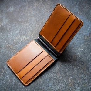Può includere: Portafoglio in pelle marrone chiaro aperto, con fermasoldi. Il portafoglio ha più fessure per carte ed è cucito con un filo più chiaro. L'interno è nero. Il portafoglio è aperto.