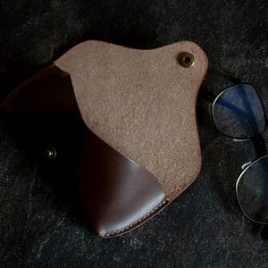 May include: A brown leather glasses case with a flap closure and a snap button. The case is open and the inside is lined with a soft, brown fabric.