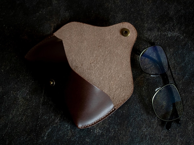 May include: A brown leather glasses case with a flap closure and a snap button. The case is open and the inside is lined with a soft, brown fabric.