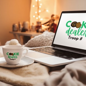Puede incluir: Un ordenador portátil blanco con un logotipo verde y marrón que dice "cookie dealer Troop #" en la pantalla. Una taza de café blanca con el mismo logotipo está en un platillo blanco delante del portátil.