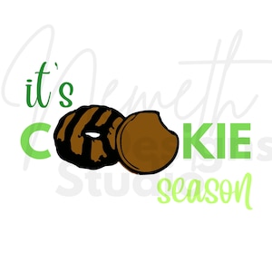 Puede incluir: ¡Es la temporada de galletas! Texto verde con una ilustración en negro y marrón de dos galletas Girl Scout.
