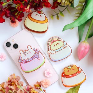 Könnte beinhalten: Ein rosa iPhone 13-Hülle mit einem Pop-Socket mit einem Cartoon-Hasen, der von Blumen umgeben ist. Zusätzliche Pop-Sockets mit Dessert-Designs, darunter ein Pudding, ein Bär auf einem Kuchen und ein Regenbogen, sind verstreut.
