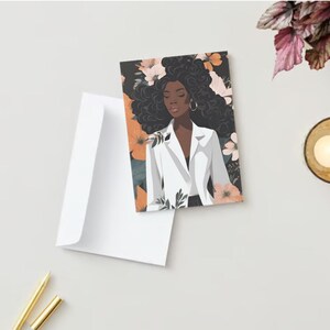 May include: Greeting card featuring an illustration of a woman in a white blazer with floral accents. The card is paired with a white envelope. A lit candle in a gold-colored holder and two gold pens are also visible.